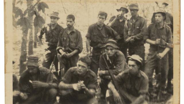 A picture from Phuoc Tuy Province, Long Tan on August 19, 1966. Informal group portrait of members of D (Delta) Company, 6RAR, taken the morning after the Battle of Long Tan. The men are listening to their Company Sergeant Major (CSM) 2/7758 Warrant Officer 2 John William 'Jack' Kirby, who is relaying a message from Australian Prime Minister, Mr Harold Holt. Identified are, back row, left to right: Barry Richardson; 15230 Corporal (Cpl) Jeffrey (Jeff) Max Duroux, an Aboriginal serviceman; 1731007 Private (Pte) Allen James May; 2781700 Pte Noel John Grimes; Ronny Moss; 1730914 Pte David John Collins. Front row, left to right: unidentified; Peter Ainsley; unidentified and unidentified. This is a film still captured from Department of Public Relations footage DPR/TV/426-427, and which is held in the Memorials collection at F10108. Picture from Australian War Memorial/P05889.001